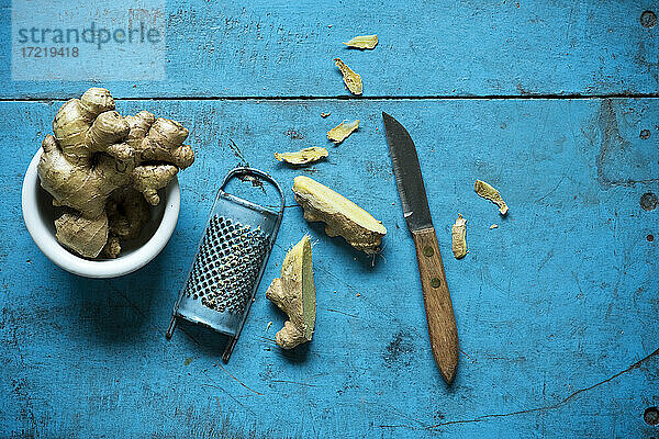 Ingwerwurzel,  Reibe und Küchenmesser liegen auf einer blauen Holzfläche