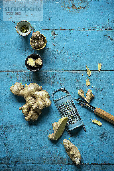 Ingwerwurzel,  Reibe,  kleine Tassen und Küchenmesser auf einer blauen Holzunterlage