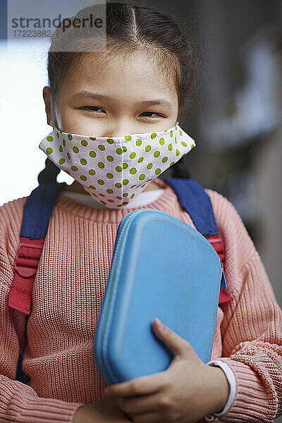 Mädchen mit Gesichtsschutzmaske,  das während einer Pandemie ein Mäppchen hält