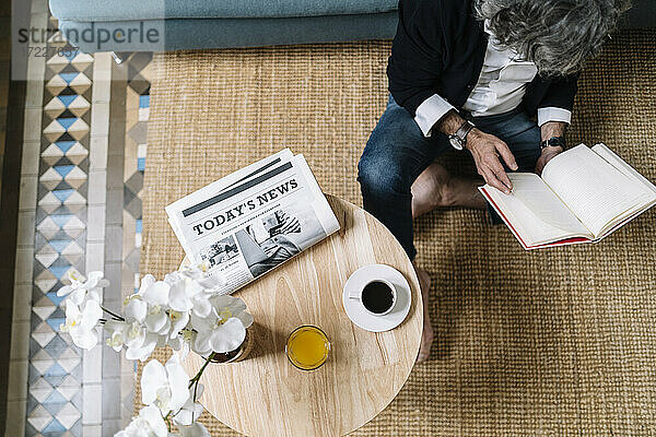 Mann,  der ein Buch liest,  während Kaffee und Saft auf einem Holztisch zu Hause stehen