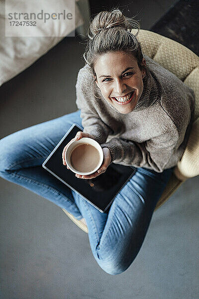 Lächelnde erwachsene Geschäftsfrau,  die eine Getränkeschale hält,  während sie mit einem digitalen Tablet in einem Café sitzt