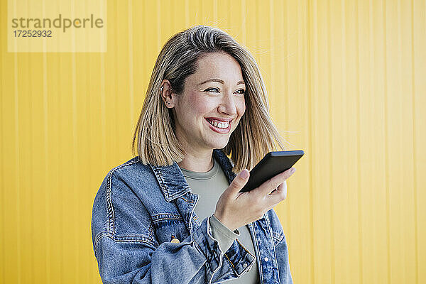 Glückliche Frau,  die eine Sprachnachricht über ihr Smartphone an eine gelbe Wand sendet