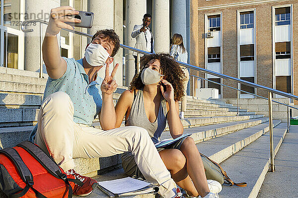Männlicher Freund gestikuliert Frieden,  während er ein Selfie mit einem Universitätsstudenten mit Schutzmaske auf einer Treppe in der Stadt macht