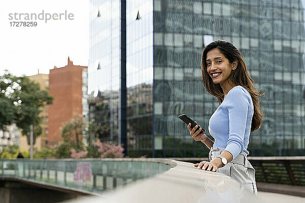 Glückliche junge Geschäftsfrau mit Mobiltelefon auf einer Fußgängerbrücke