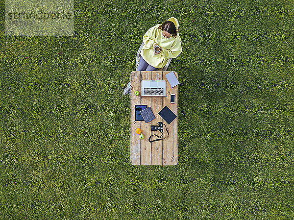 Luftaufnahme einer Frau,  die an einem Kaffeetisch auf grünem Rasen sitzt und eine Tasse Kaffee in den Händen hält