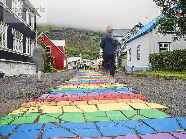 Island,  Seydisfjordur,  Menschen gehen entlang der regenbogenfarbenen Asphaltstraße inmitten einer abgelegenen Stadt
