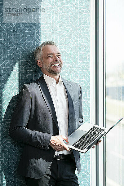 Glücklicher Geschäftsmann hält Laptop vor der Wand im Büro