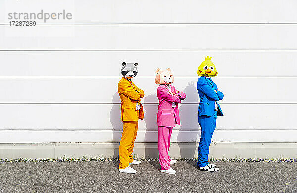 Drei Personen in farbenfrohen Anzügen und Tiermasken posieren nebeneinander vor einer weißen Wand