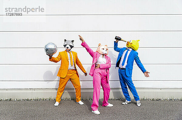 Drei Menschen in farbenfrohen Anzügen und Tiermasken feiern vor einer weißen Wand