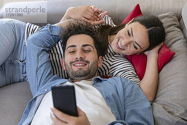 Glückliches Paar,  das ein Mobiltelefon benutzt,  während es zu Hause auf dem Sofa liegt