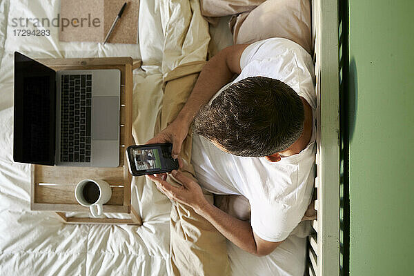 Ein Mann führt einen Videochat mit einem Freund über sein Mobiltelefon,  während er im Bett am Laptop sitzt