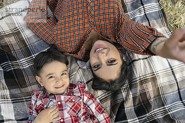 Lächelnde Mutter,  die ein Selfie mit ihrem Sohn macht,  während sie auf einer Picknick-Decke liegt