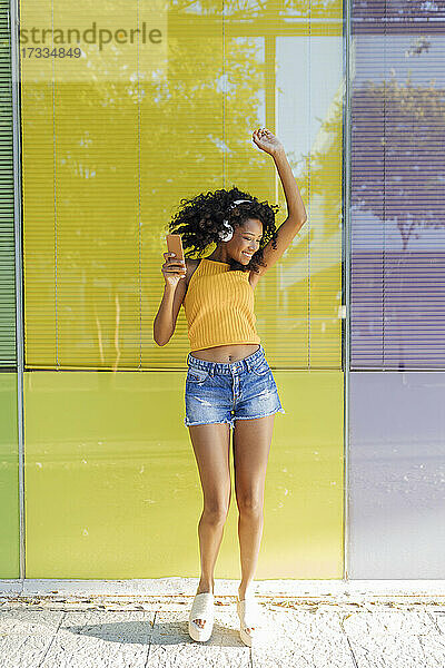 Glückliche Frau mit erhobener Hand tanzt vor einem gelben Fenster