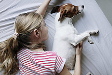 Blondes Mädchen liegt auf Bett und kitzelt ihren Hund