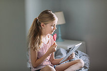 Blondes Mädchen sitzt mit Lolli auf dem Bett und benutzt ein digitales Tablett